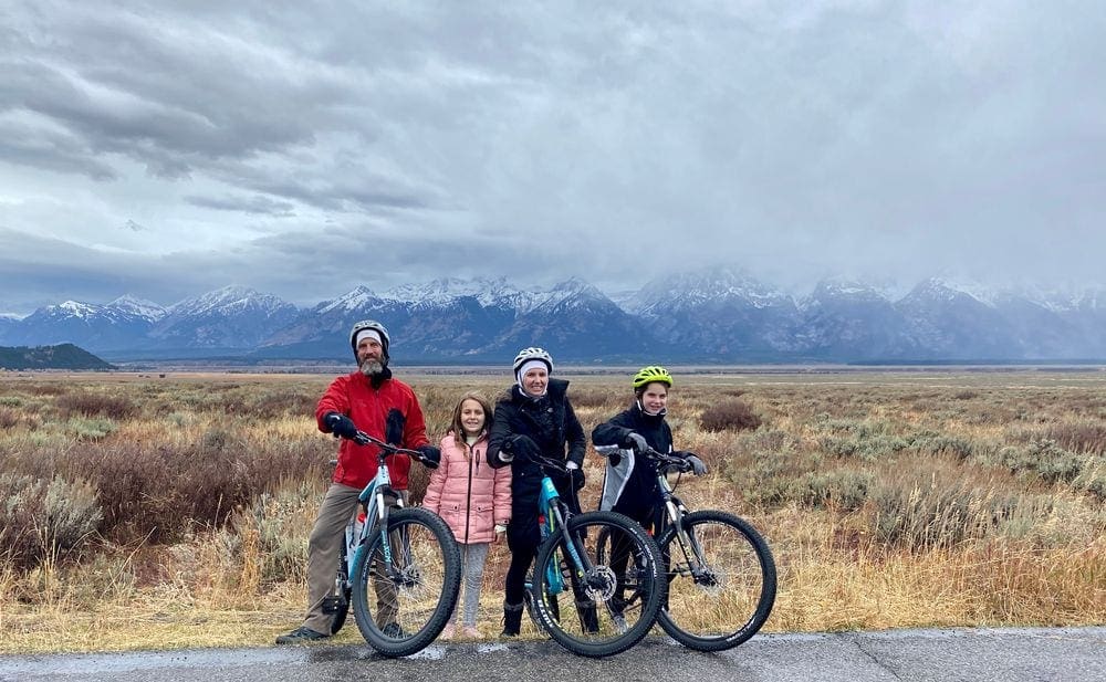 A family of four wearing coats and helemets pose togehter while taking a break from biking, one of the best things to do in Grand Tetons National Park with kids.