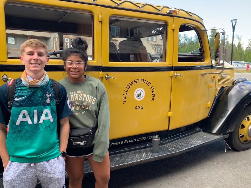 Two tweens standing in front of the yellow Yellowstone National Park shuttle.