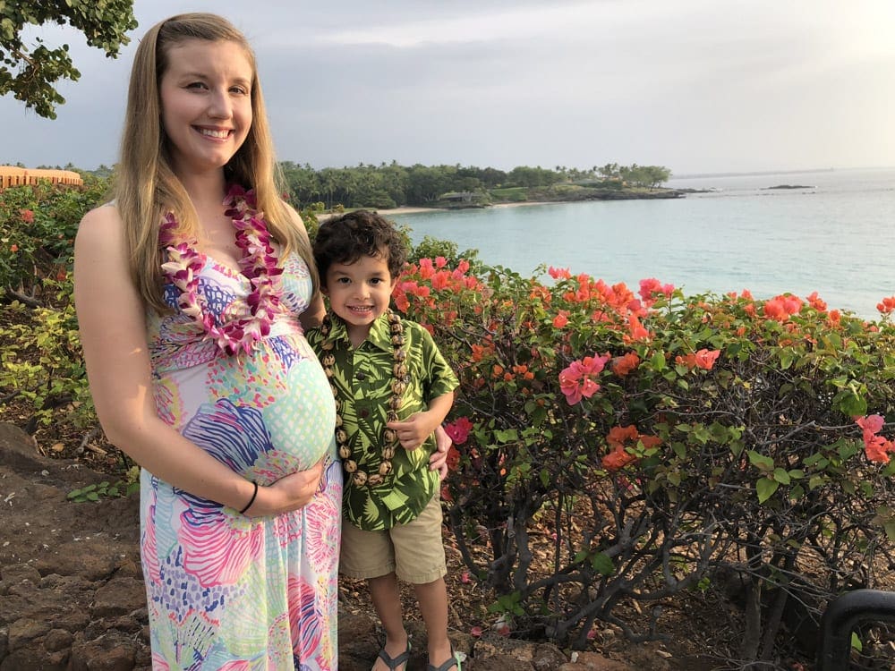 A pregnant mom holds her tummy, while standing next to her toddler son on a beautiful day along the water in Maui.