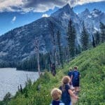 Four kids hike along a lush path with the Grand Tetons in the distance.