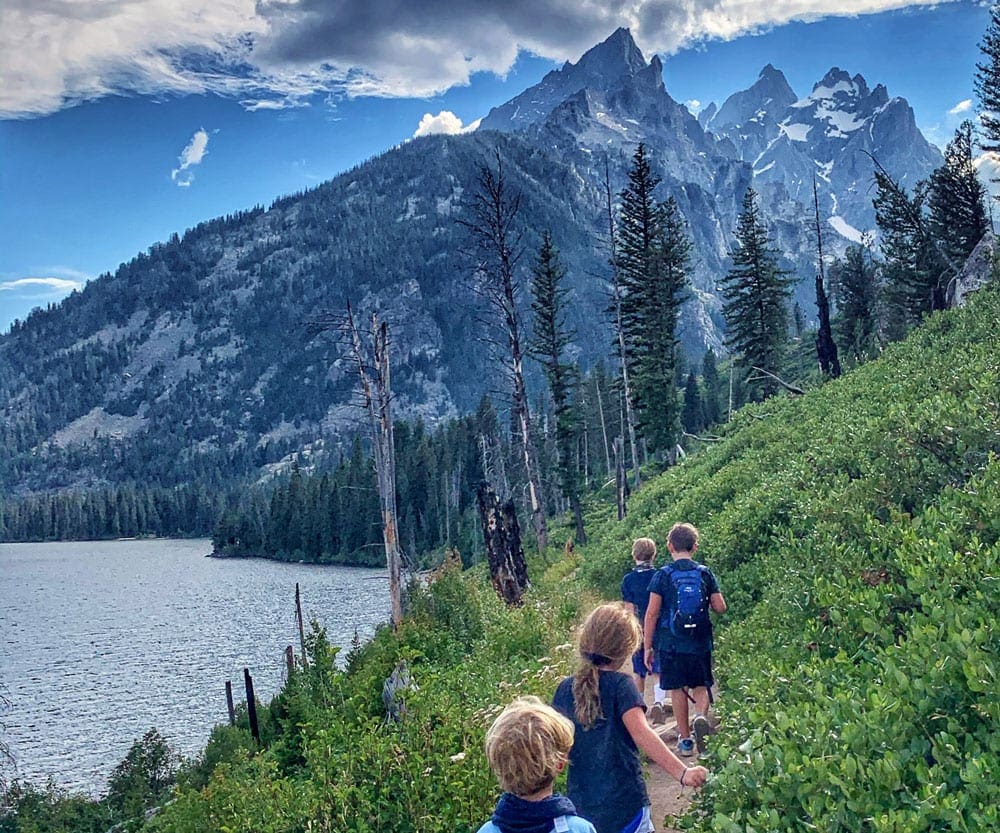 Four kids hike along a lush path with the Grand Tetons in the distance.