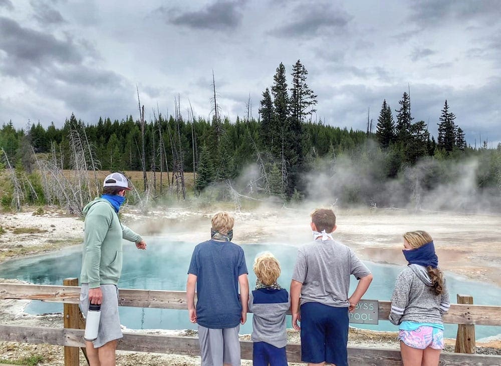 A family of five peers into the teal hot spring at Yellowstone National Park. It's one of the best places to visit on a Yellowstone itinerary for families!