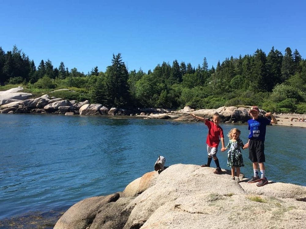 Three kids stand hand in hand while posing on a large boulder while hiking in Acadia National Park, one of the best places for an East Coast road trip for families. 