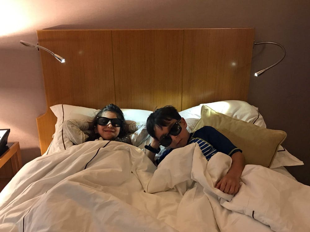 Two kids wearing sunglasses smile while snuggled in bed at The Colonnade Hotel.