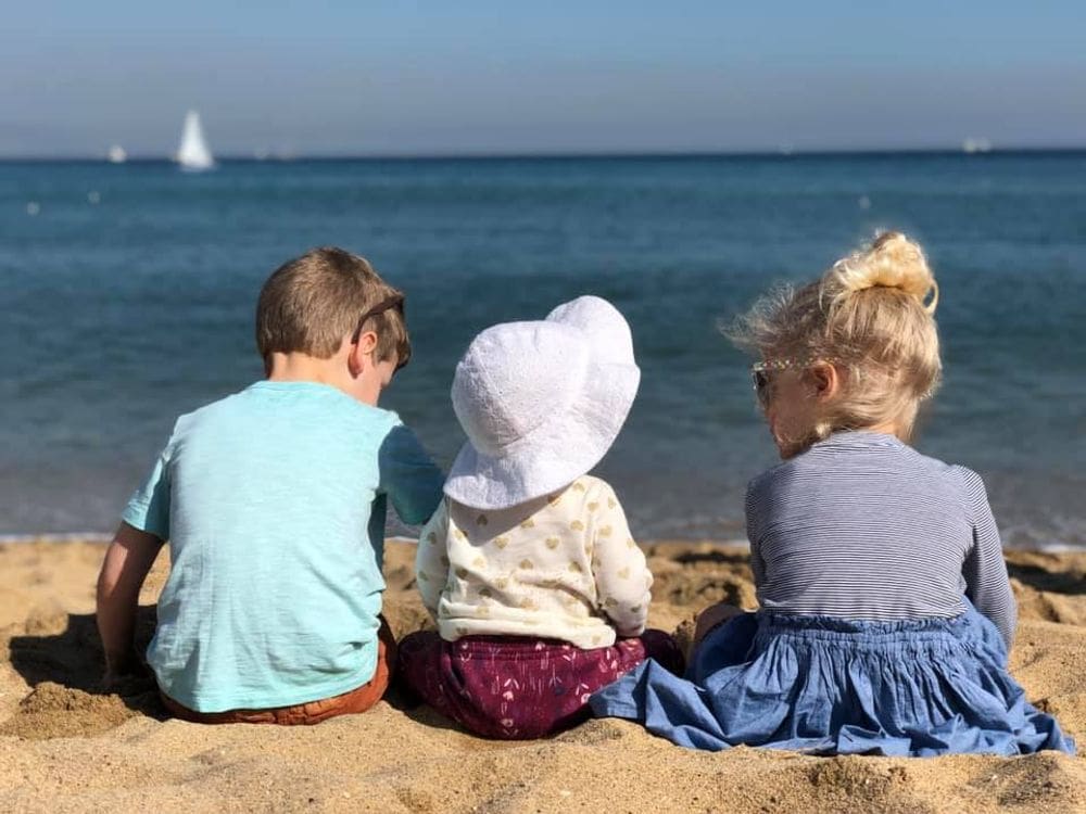 Three kids sit on the beach in Barcelona, Spain, with the water in the distance.