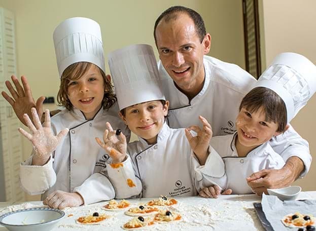 Three kids with chef hats on pose with a chef leading a kids club cooking class at the Constance Moofushi Maldives, one of the best family hotels in the Maldives.