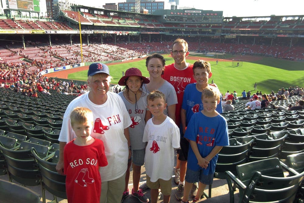 A family of eight poses together with Fenway Park in the background as they enjoy a baseball game on a sunny day.