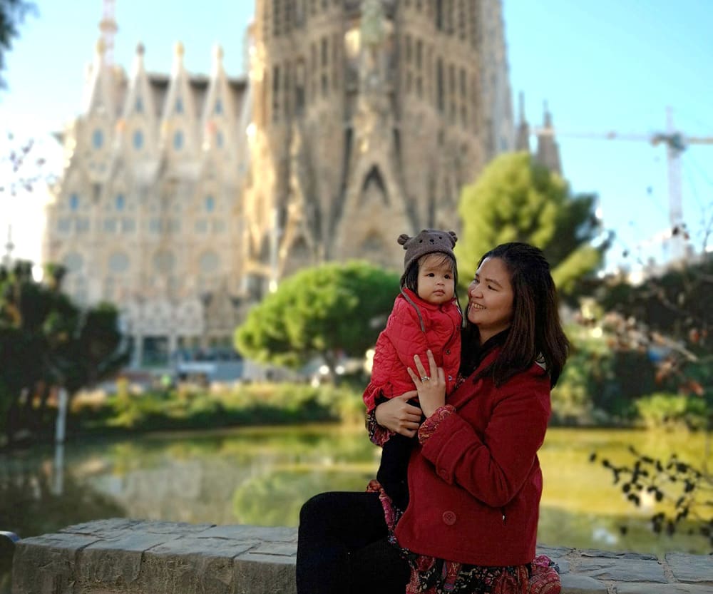 A mom holds her young son while exploring Barcelona, Spain, with La Sagrada Familia in the distance.