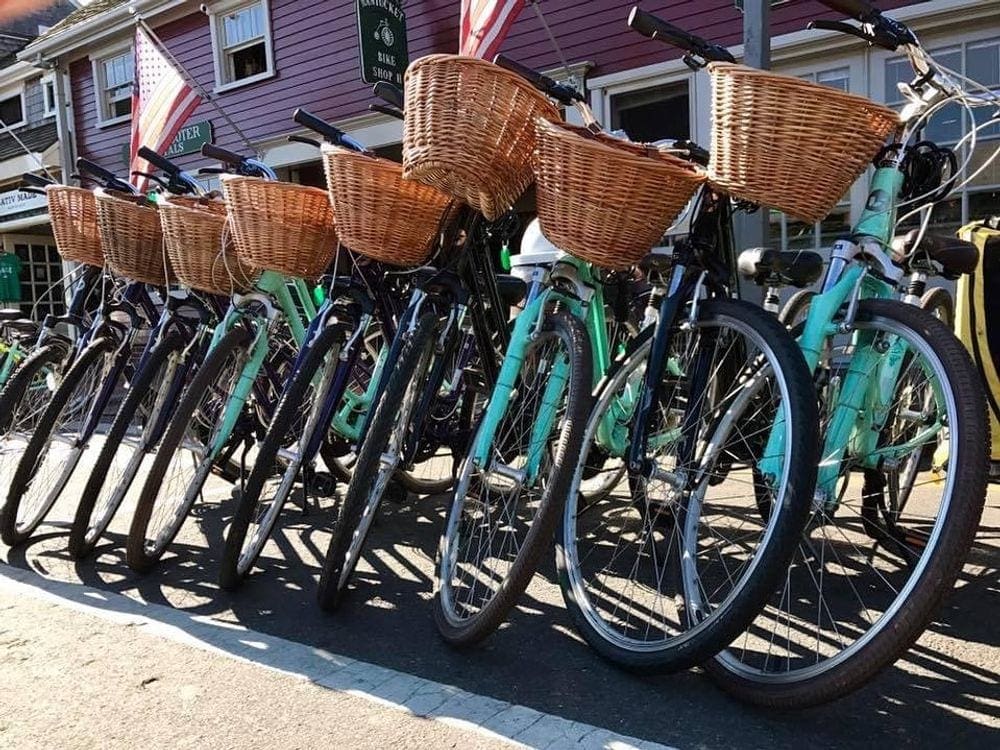 Eight green bikes with large wicker baskets rest on their stands waiting for a tour during a family vacation to Nantucket..