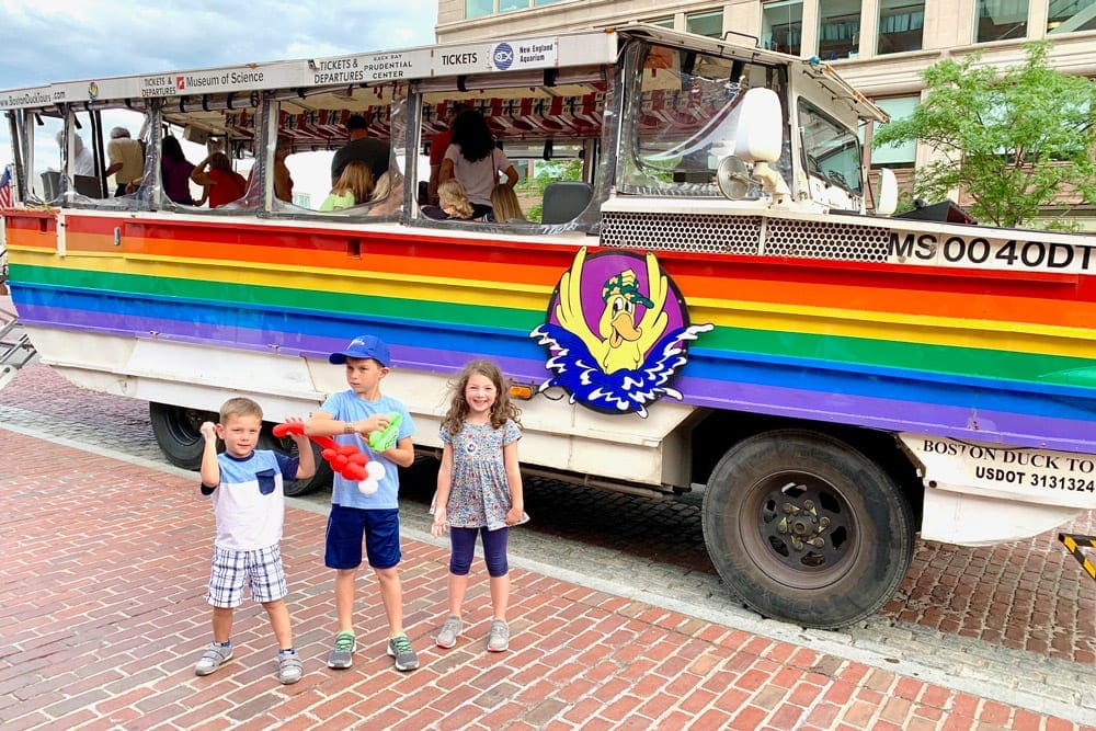 Three kids stand ashore in front of the Boston Duck Tour boat.