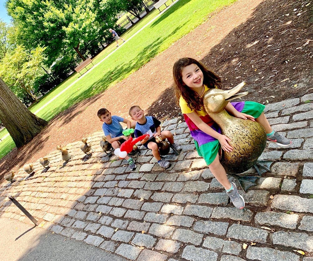 Three kids sit amongst the iconic statue of Ducks in Boston, one of the best US cities for a Memorial Day Weekend with kids.