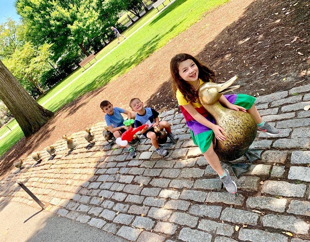 Three kids sit amongst the iconic statue of Ducks in Boston, one of the best places to visit during Memorial Day Weekend near NYC for families.