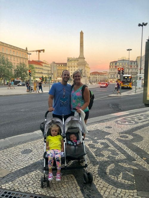 Two parents hold a double stroller, where their two kids sit, with a view of Lisbon behind them.