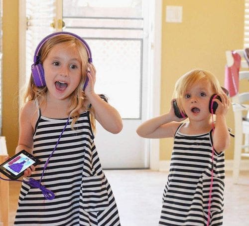 Two young girls wear the AILIHEN I35 Kid Headphones, one set is pink and one set is purple, while they dance about the house, one of the best gifts for travelers.