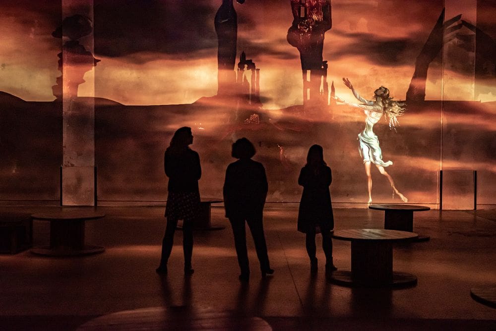 Three individuals stand in silhouette looking at an art installation inside the Atelier des Lumières.