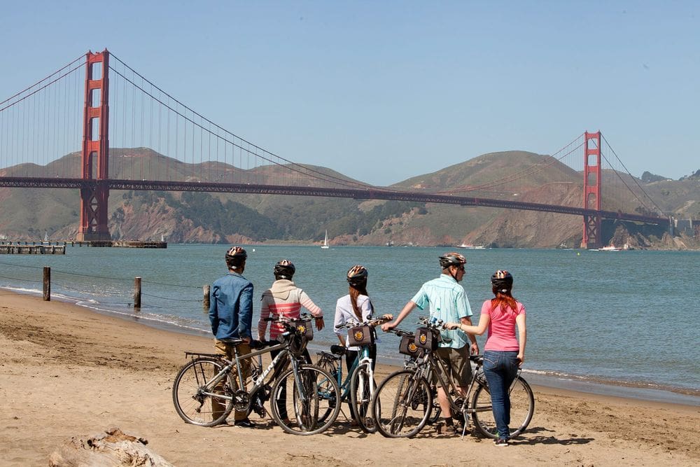 Five bikers stand with their bikes on the beach with the Golden Gate Bridge in the distance.