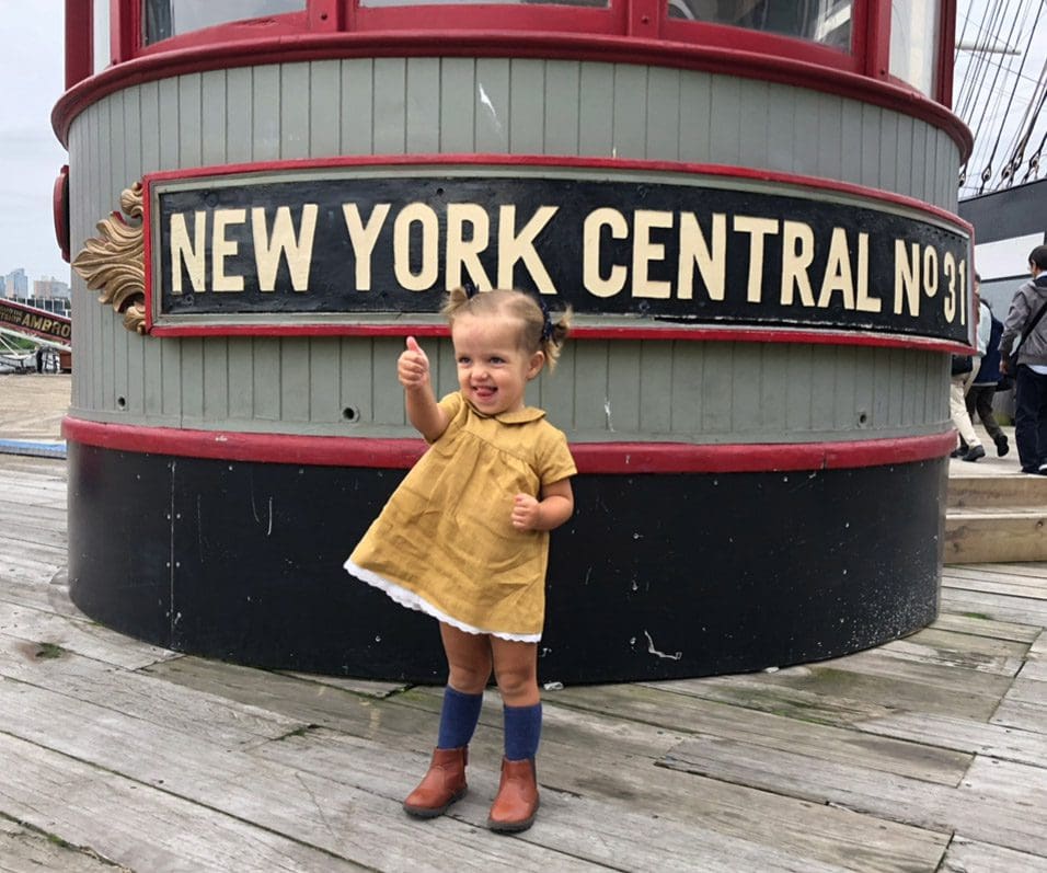 A young girl in a yellow dress gives a thumbs up outside the New York Central harbor, one of the best places to visit with your young daughter in America.