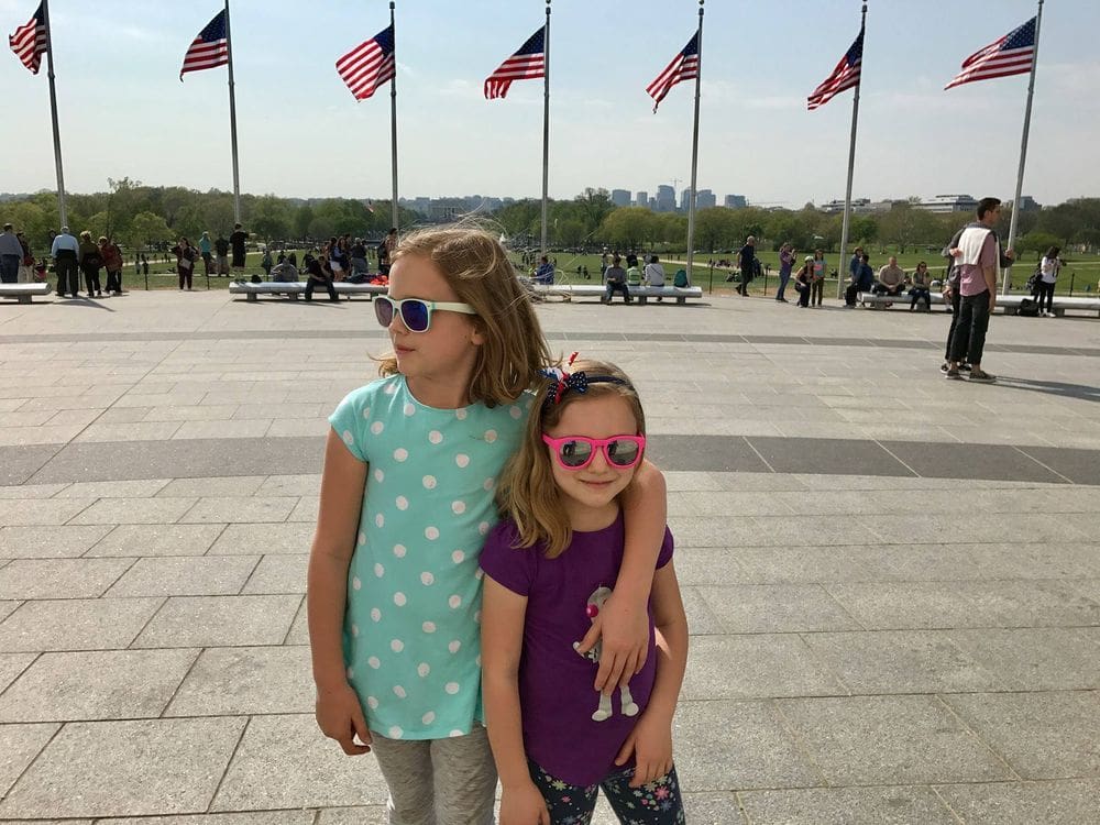 Two girls, wearing sun glasses, stand together with several American flags in the background in Washington DC, one of the best places to visit during Memorial Day Weekend near NYC for families.