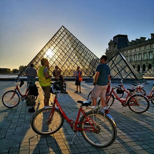 A tour group of people on bikes passes the Lourve at sunset.
