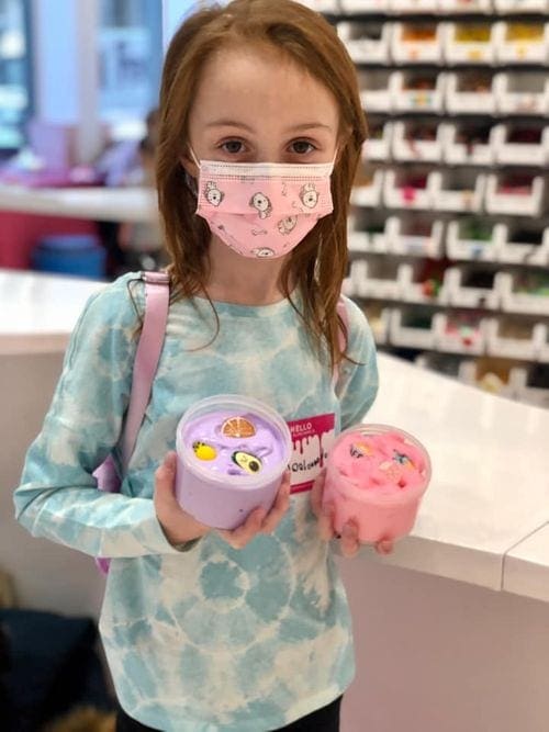 A young girl with red hair holds two containers of slime, one pink and one purple, at the Sloomoo Institute.