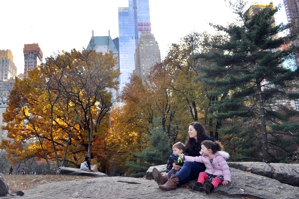 A mom and two kids sit on a large rock surrounded by fall foliage while exploring Central Park.