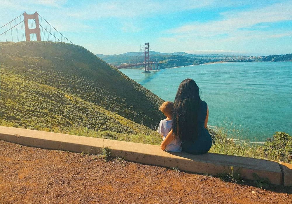 A mom and her young son sit together amongst green grass with the iconic Golden Gate Bridge in the distance.