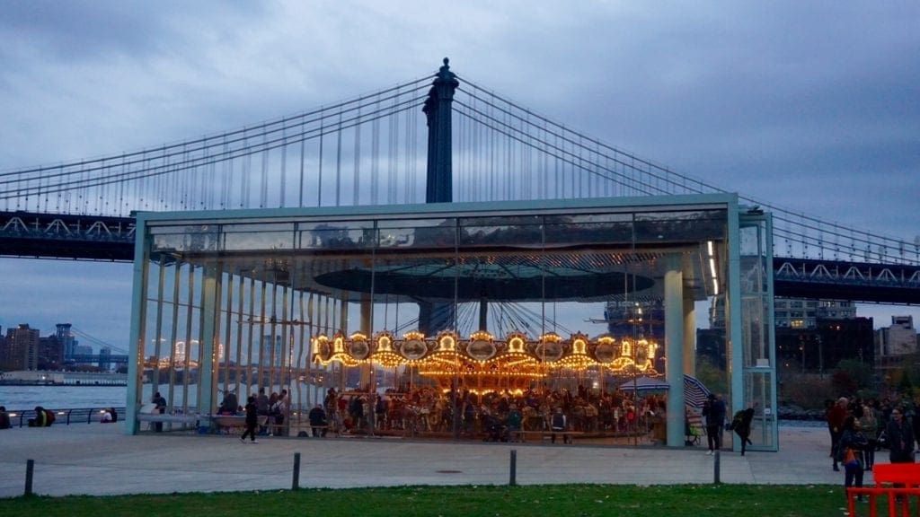 A brightly lit carousel with the Brooklyn Bridge Park in the distance.