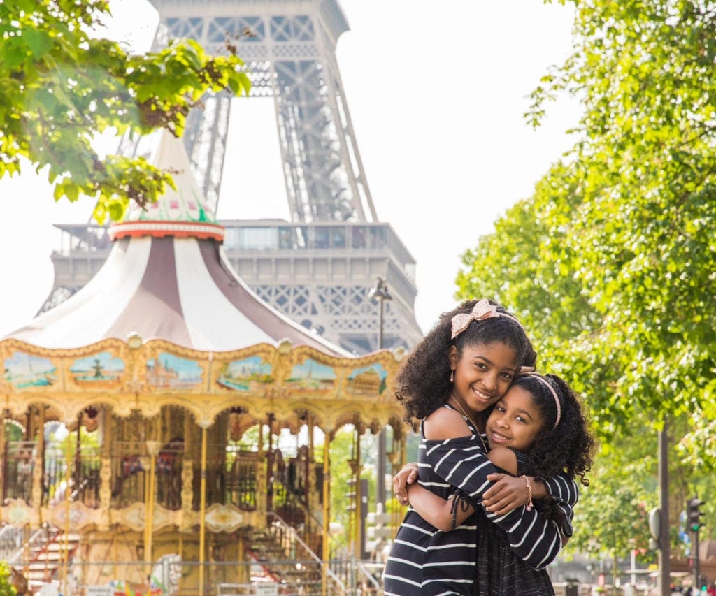 Two sisters of color embrace with Paris' iconic carousel and Eiffel tower behind them, one of the best places to visit in France with kids.
