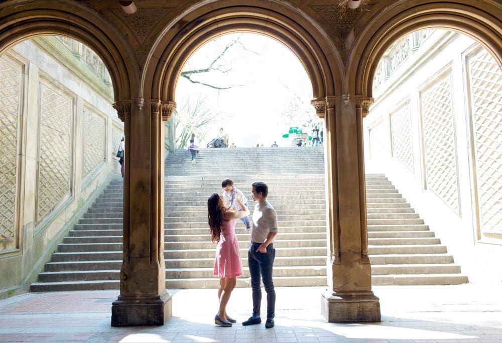 Two parents hold up their child with an adoring look when enter the iconic arches of Central Park in NYC, one of the best American cities that feel like Europe for families.