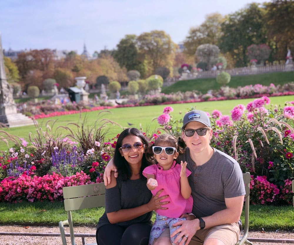 A family of three sits together with a stunning Parisian garden beyond them.