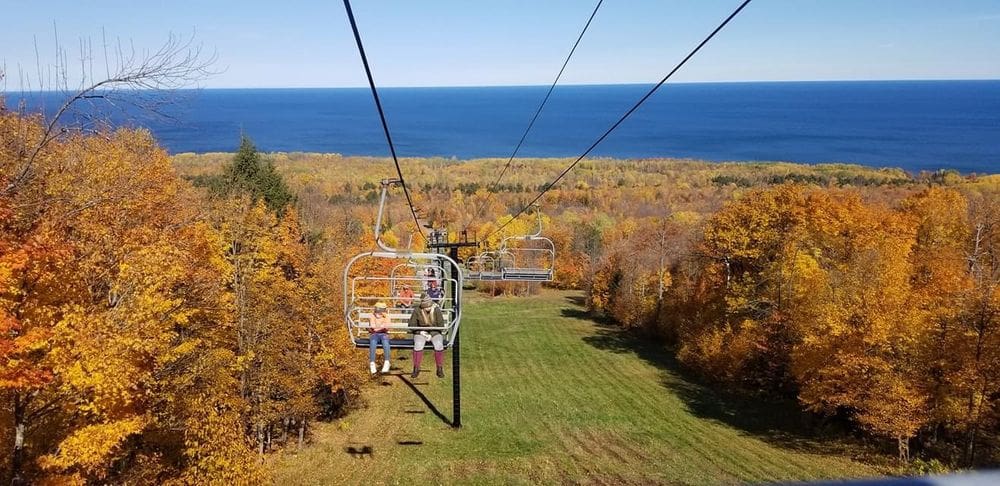 Two people ride on a chair lift up the Porkies with a view of brilliant fall colors and Lake Michigan behind them.