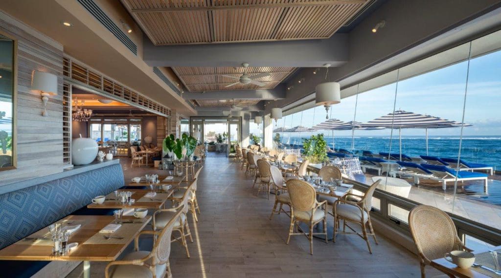 Photo of the indoor restaurant at the Condado Vanderbilt Hotel with multiple tables and blue cushion booths, overlooking the ocean.