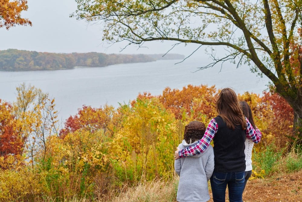 A mom puts her arms around her children, one on each side, as they look out onto a beautiful autumn view of Lake Onalaska, near La Crosse, one of the best places to see fall colors in the Midwest for families.