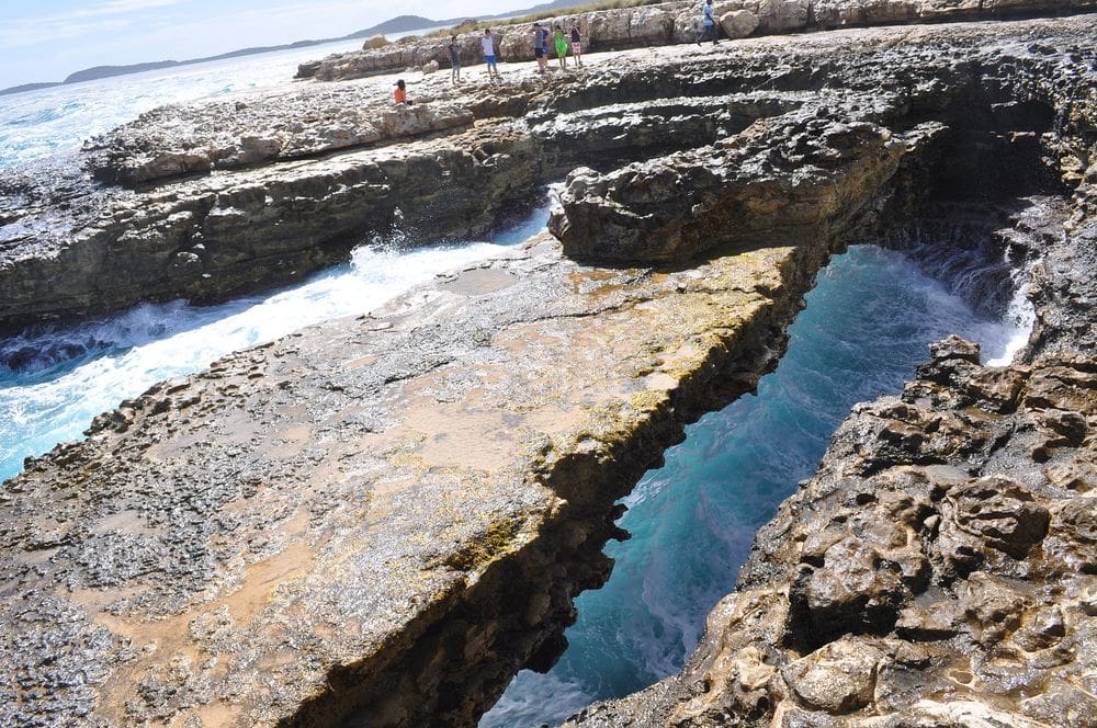 Devil's Bridge in Antigua, featuring large stones, where people are milling about, as well as ocean waves crashing against the rocks.