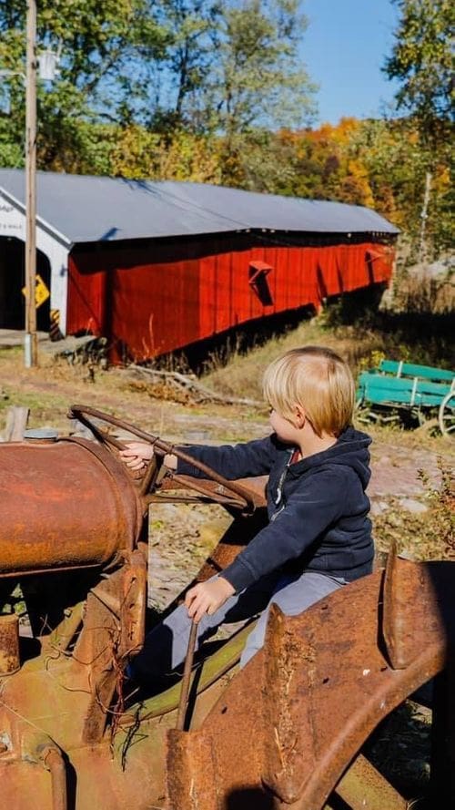 A young boy sits on a rusted tractor looking at a red covered bridge in Parke County, Indiana, one of the best places to see fall colors in the Midwest for families.