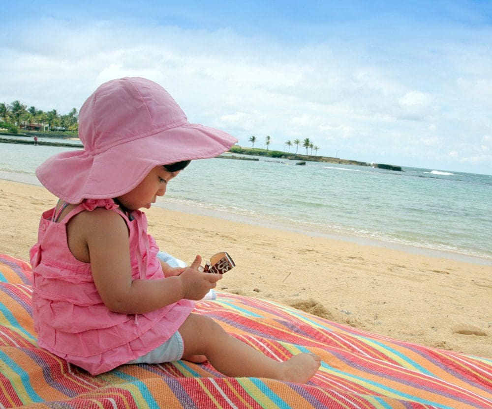 An infant girl, wearing a pink outfit and pink beach hat, sits on a vibrantly colored blanket on the beach in Puerto Rico.