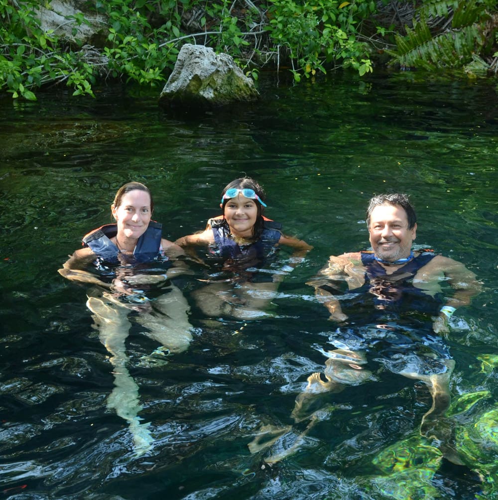 A family of three, all wearing life jackets, floats in a Cristalino Cenote, one of the best day trips from Playa del Carmen for families.