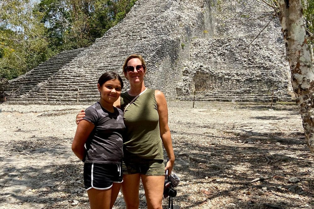 A mom and daughter stand together with a Mayan pyramid behind them, located near Coba.