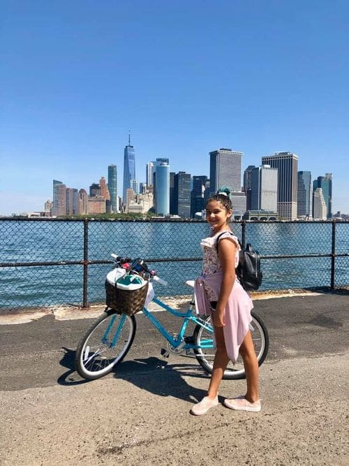 A young girl poses in front of her bike while exploring Governor's Island on a sunny day, the NYC skyline is seen in the distance.