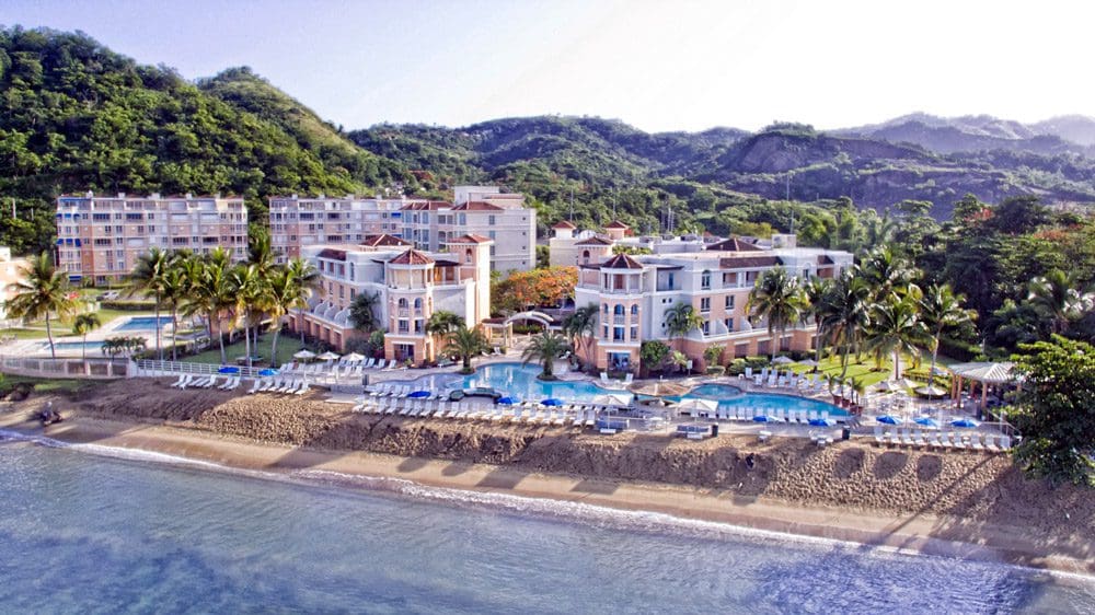 Picture of the shoreline where the Rincon Beach Resort is located, featuring a large outdoor pool and stretch of beach. The hotel's signature peach-colored buildings stand in the background.