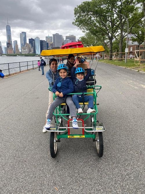 A family of four enjoys a surrey bike around Governor's Island with the NYC skyline in the distance.