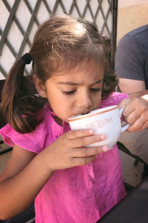 A young girl with a pink shirt and pigtails in her hair drinks from a delicate tea cup reading "Angelina", one of the best dessert destinations in Paris for families.