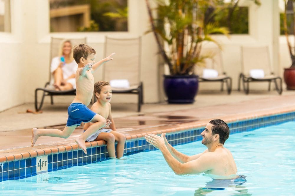 A young boy jumps into his dads arms, who is standing in the pool at the Bahia Resort Hotel, while mom and brother look on at one of the best San Diego beachfront hotels for families.