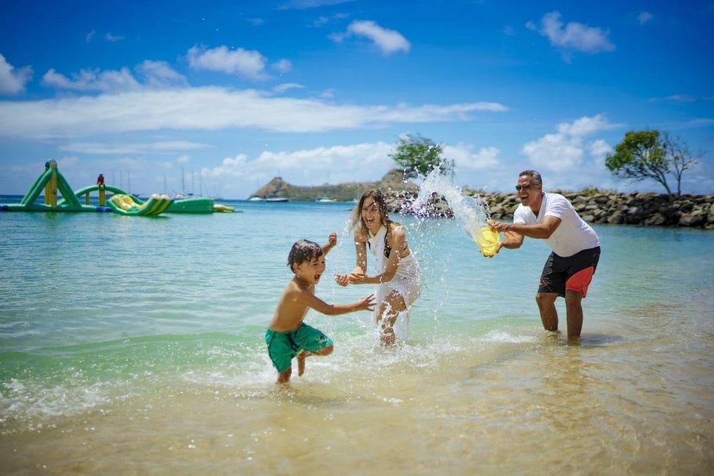 A dad splashes water at the mom and child while playing off-shore from Bay Gardens Beach Resort & Spa, in the distance large inflatables can be seen.