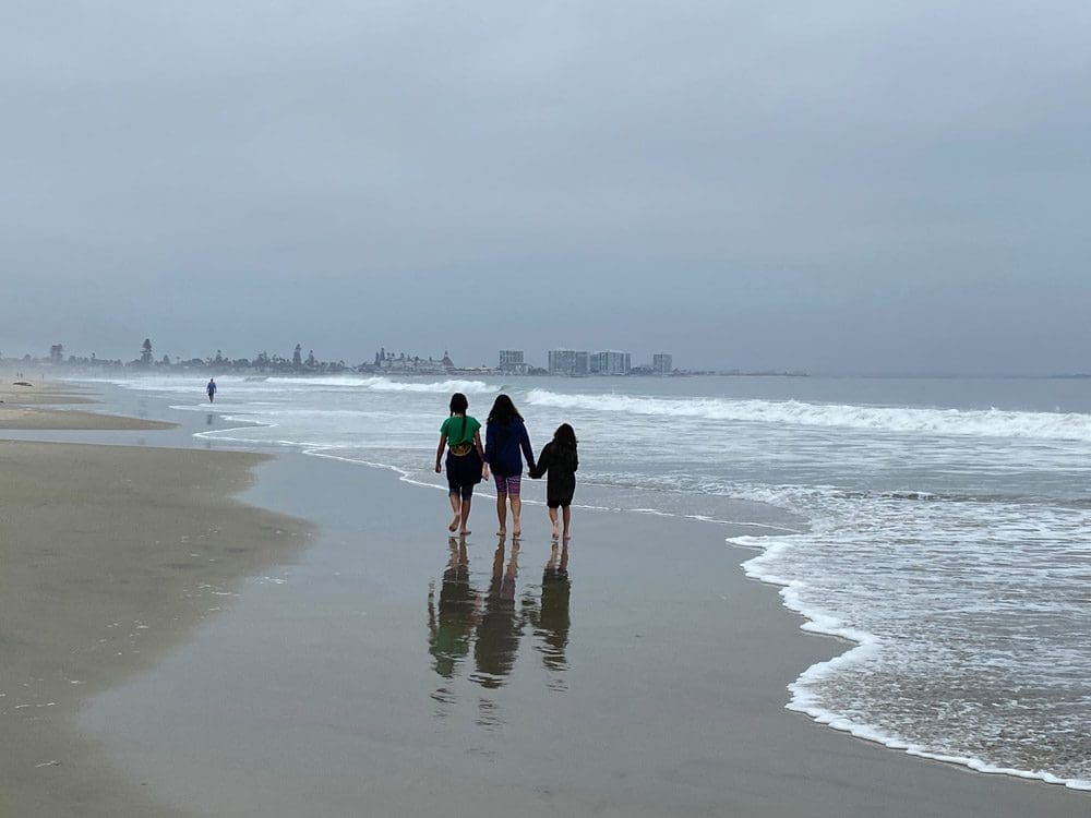 A mom with two daughters, one on each side of her, walks down a beach in San Diego, California on an overcast day.