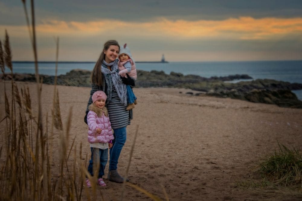 A mom stands next to one daughter, while holding another, on an autumn day on a beach in West Have, Connecticut. A lighthouse is seen in the distance.