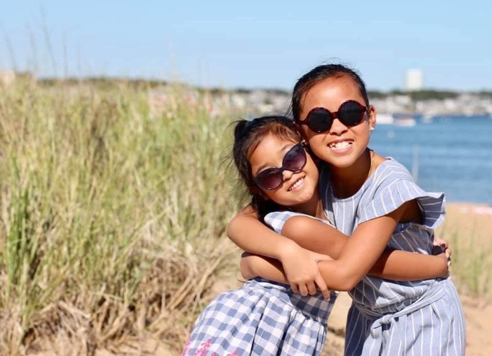 Two sisters wearing lovely dresses hug one another while enjoying a sunny day on the beach in Cape Cod, one of the best Labor Day Weekend getaways near NYC with kids.