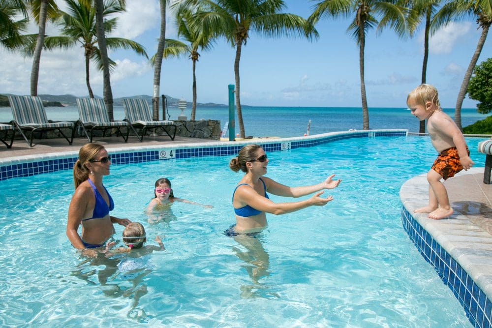 Two moms and three kids play in the pool at The Buccaneer Hotel, one child is on the pool deck ready to jump into the arms of one of the moms while the others look on.