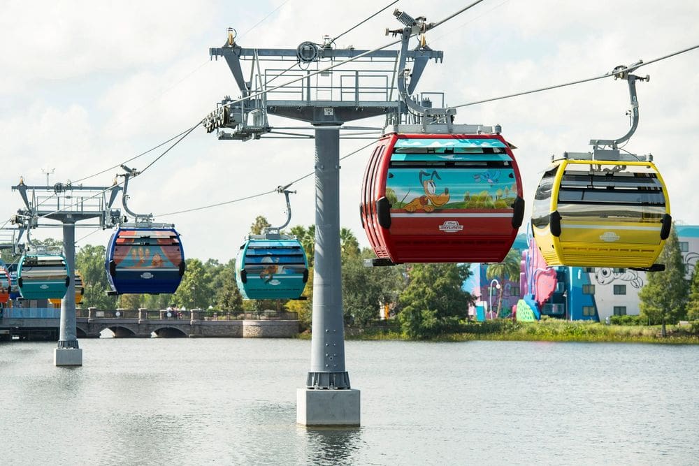 Several character-themed cars above the water move forward on the Disney Skyliner.