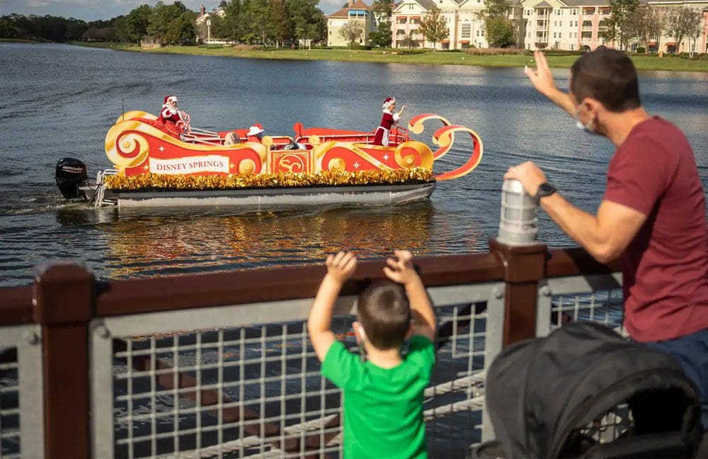 A father and son standing on a bridge wave to Santa and Mrs. Clause as they pass by on a holiday float near Disney Springs.